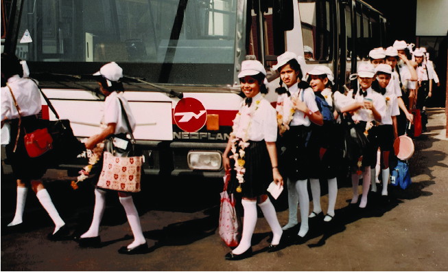 Children in uniform, wearing garlands and carrying bags, boarding a coach