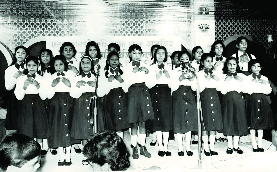 Two rows of girls in white blouses and dark skirts singing at microphones, beneath a banner naming Kochi Konther Asar