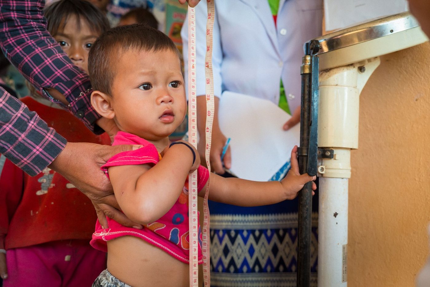 A young child standing against a height board while a health worker takes the measurement. A child measured against a height board at a rural clinic, Laos.