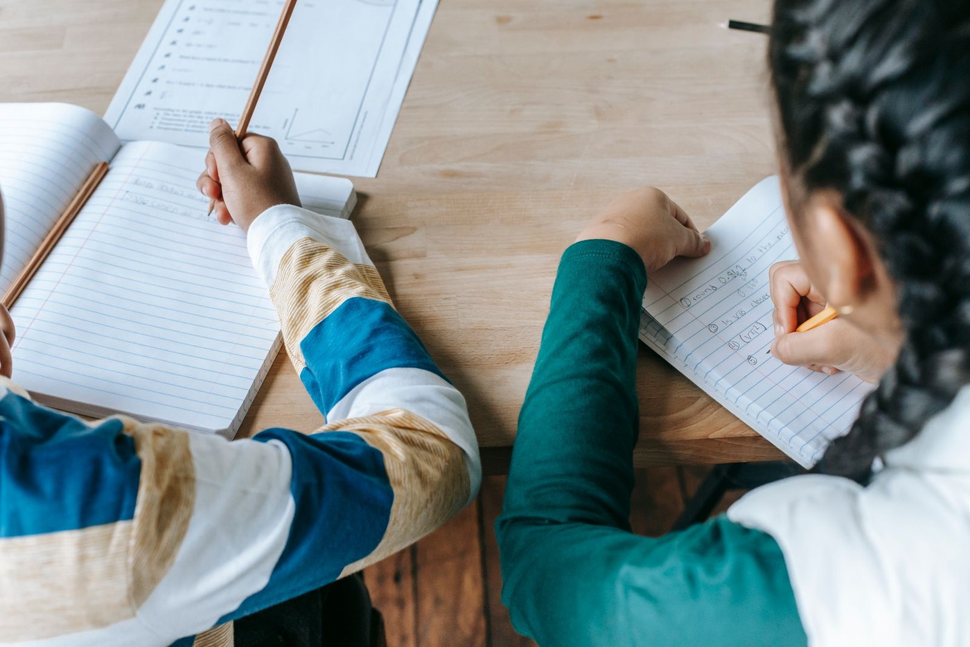 Two children writing in exercise books at a table, seen from above; neither child’s face is visible